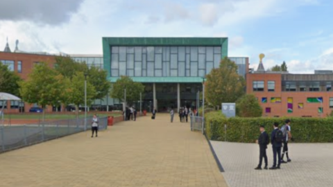 General view of Highfields School in Wolverhampton, a red brick building with a green rectangular section at the front with windows. A handful of children with blurred out faces can be seen near the entrance