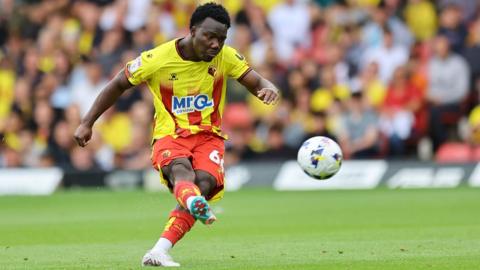 Nestory Irankunda of Watford kicks a ball off the ground on the football pitch with a focused look onhis face. he wear a yellow and red striped top with red shorts.ts.