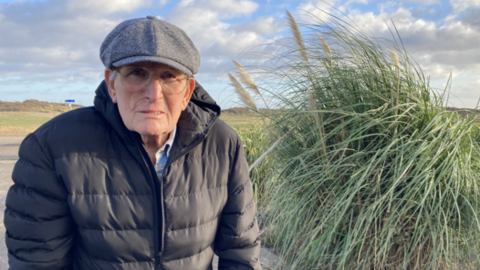 An elderly man wearing a cap and glasses with a black jacket looking directly into the camera. On the right is a green read bush.