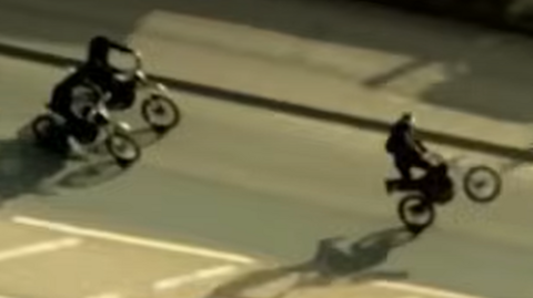 Three people on bikes, in a road, the image is taken from a plane so slightly blurred. One person looks like they are doing a wheelie.