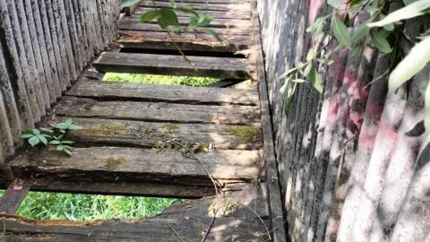 A footbridge that is falling apart. Some wooden slats are missing and you can see the ground beneath the bridge.