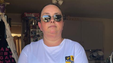 A woman with dark brown hair slicked back from her face, standing in a kitchen doorway wearing a white t-shirt, sunglasses and holding a walking stick for the blind.