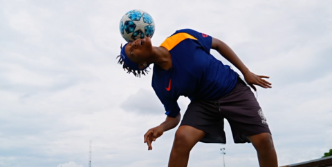 A camera shoots up from a low level to show a female freestyler leaning to the side while balancing a football on her head underneath a cloudy sky