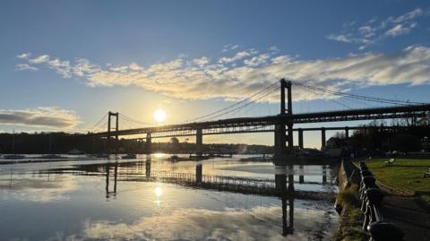 The Tamar Bridge over the River Tamar in Devon and Cornwall. The sun is seen setting behind the bridge and it is reflected on the water.