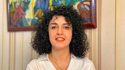 A headshot of Narges Mohammadi  - she looks towards the camera and smiles while wearing a white blouse.