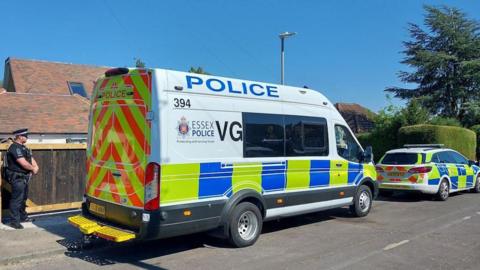 A police car and van parked on a residential road, which has a large tree, bush and house on it. A police officer guards the entrance to a property.