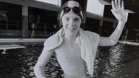 Black and white photograph of a boy standing next to an indoor swimming pool with a swimming cap and goggles on his head and a towel over his shoulders