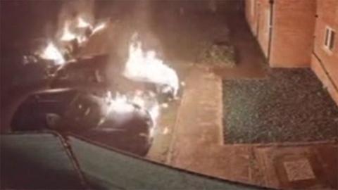 CCTV still showing a number of cars on fire outside modern-looking terraced houses