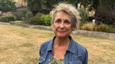 Veronica Dewan standing on a grassy area outside Parliament. She has a blue denim jacket on and a blue headband over white blonde hair.