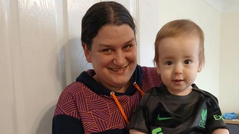 Emma with dark hair and a purple and black hooded top sits with Jack on her knee. He has brown hair and is wearing a black and green Liverpool FC football shirt