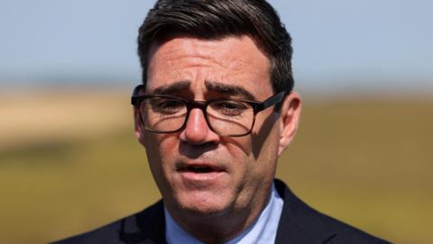 Andy Burnham looking thoughtful against backdrop of a field. He is wearing dark rimmed spectacles