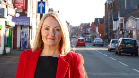 Portrait of MP Jo Platt standing on Railway Road, Leigh. She is wearing a red jacket and smiling.