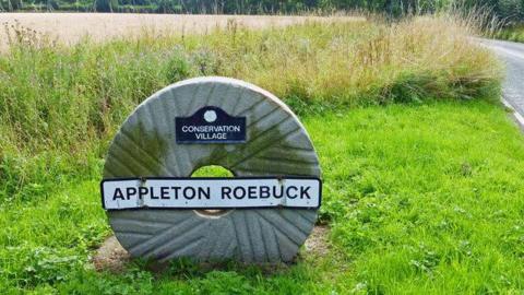 A stone circle with a hole in the middle on a grass verge with the words Appleton Roebuck written on it.
