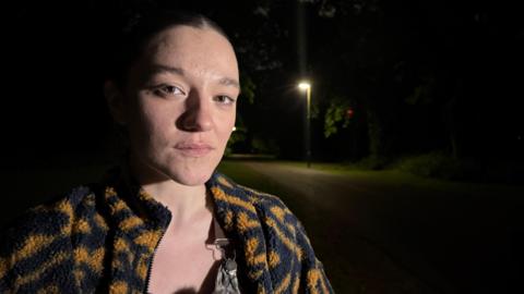 A woman stood looking at the camera with a street light in the background which is next to a green bush.