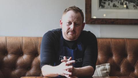 Kenny Atkinson sitting on a brown leather banquette looking into the camera with a straight face. He has his hands clasped in front of him on a table and he is leaning forward slightly. He has brown, short hair which is cropped to the side and a beard. He is wearing a black long-sleeved top which is rolled up and a blue House of Tides apron.