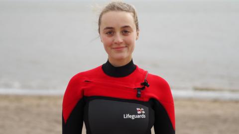 Lois Kemp stood on a beach, smiling at the camera. She is wearing a black and red swimsuit with the RNLI Lifeguards logo on her chest. She has blonde hair tied back. 