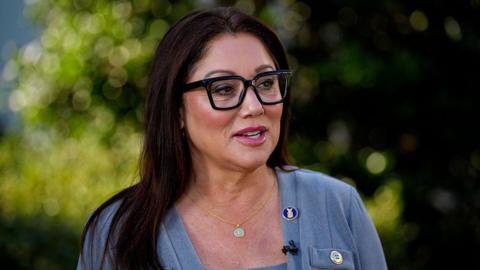 Lori Chavez-Deremer in grey sweater, gold necklace, and black glasses