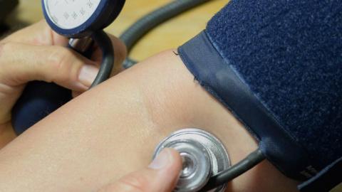 A stock image of a patient getting their blood pressure measured. The image is a close-up showing the hands of the medical professional holding the blood pressure measuring equipment on the patient's arm.