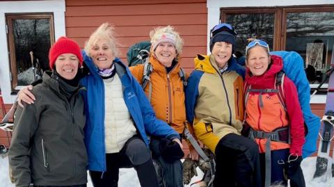 Five women in ski gear smiling to camera hugging each other