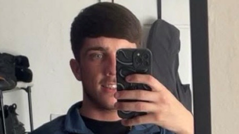 Jordan Orrock is smiling in the mirror for a selfie. He has short, but full, brown hair in a bowl cut and is wearing a blue shirt. He has short, brown facial hair and is holding his black phone in a way which obscures half of his face.