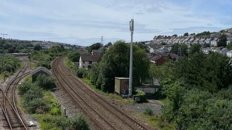 Overlooking a railway track. The train tracks are surrounded by bushes, trees and grass. Homes are visible in the distance. 