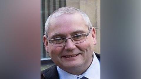 David Daintree smiles at the camera in a suit and shirt and tie. He has short grey hair and silver rimmed glasses. 