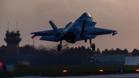 A fighter jet takes off from RAF Lakenheath. The light is low and heat coming from the jet can be seen rippling in the area. Other aviation buildings on the site can be seen in the distance.