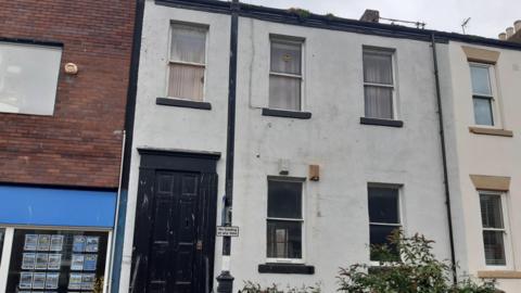 Terraced two-storey house with black door and grey paint on its walls. It has three windows on the top floor and two on the ground floor.