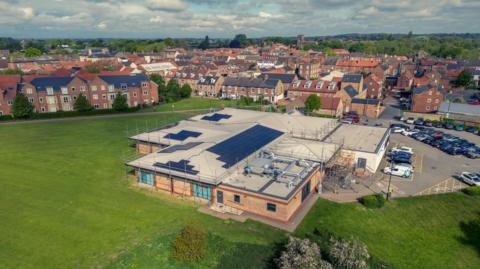 An aerial view of Thirsk and Sowerby Leisure and Wellbeing Hub, with solar panels installed on its roof. It's a large building with a mix of standard and flat roofing. There's a car park to the right of it, and a large expanse of grass around it with some trees. In the background there are residential streets, stretching off to trees and hills in the horizon. The sky is blue and cloudy.
