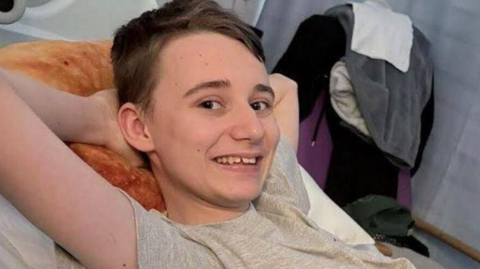 A boy with short brown hair and a grey t-shirt with his arms behind his head