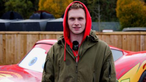 A man looking into the camera, stood in front of a bright red car. He is wearing a red hoodie and a dark green coat, and has a wireless microphone attached to his collar.