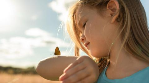 Close up shot of a young girl with long fair hair looking at a butterfly which has landed on her arm. The background shows a blurred sunny sky.
