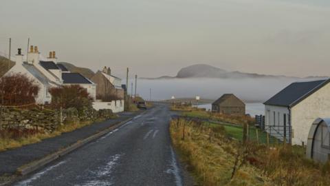 A road through a community of white-walled homes in Lewis. In the distance the top of a hills is visible behind a bank of fog.
