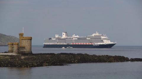 The Holland America Line’s Rotterdam, which is blue and white, out at sea in Douglas Bay. Tower of Refuge, which looks like a small castle and is on an islet in the sea, can be seen in the foreground.