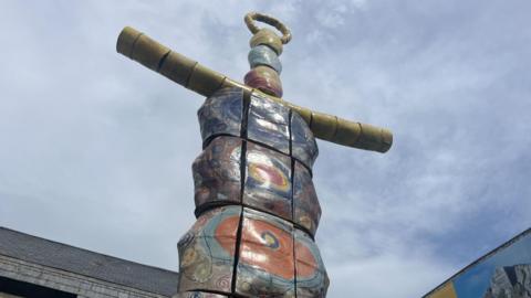 The image shows the top of a very tall statue called the Earth Goddess. It resembles a brightly coloured totem pole and has yellow arms. The sky behind it is blue with thin white cloud. The tops of two buildings are visible too.