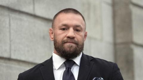 McGregor is wearing a dark grey suit and tie. He has a shaved head.