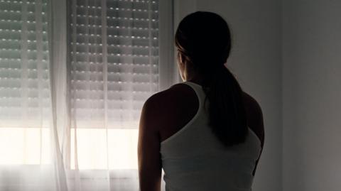 A woman with her back to the camera sits on a bed looking towards a window