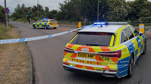 Two police cars parked either side of blue and white police tape on a main road, which is lined with trees.