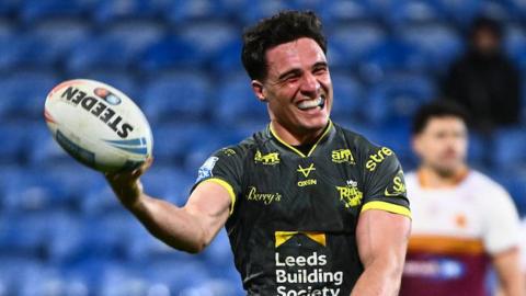 Leeds Rhinos half-back Brodie Croft holds up the ball in one hand and grins to celebrate one of his four tries against Huddersfield Giants