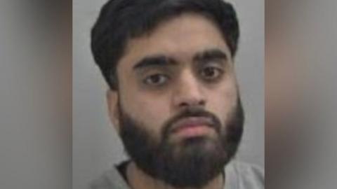 Anayat Ur-Rehman is seen in a police mugshot. He has a black beard and moustache and black hair.