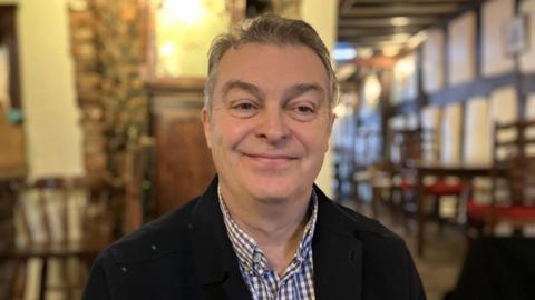 A man wearing a black jacket and a patterned shirt. He has short grey hair, and is stood in a pub.