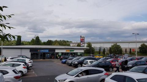 Cars parked in a retail park with Greggs and Subway seen in the background.