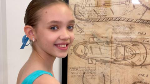 Jessica is wearing a blue leotard and is standing in front of a drawn diagram of a ballet shoe, which is stuck on a white wall. She is turn her head over her shoulder, smiling, looking at the camera. 