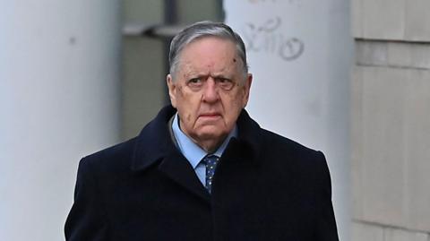 William Lloyd-Lavery walking outside. He is wearing a navy jacket over a light blue shirt with a navy tie with multi-coloured spots on it. He has grey hair. 