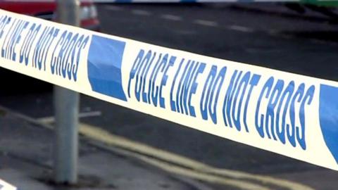 Blue and white police tape with a police officer in a hi-vis jacket over their uniform blurred in the background.