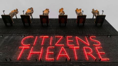 A photo of the outside of the theatre. The name of the theatre is written in red neon. Above that are the six statues which have been restored.