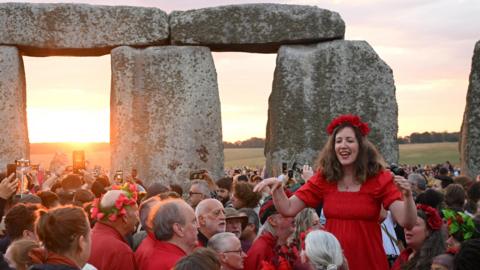 A woman in a red dress stands higher than a crowd of people within the Stonehenge stone circle. The sun is rising in the background.