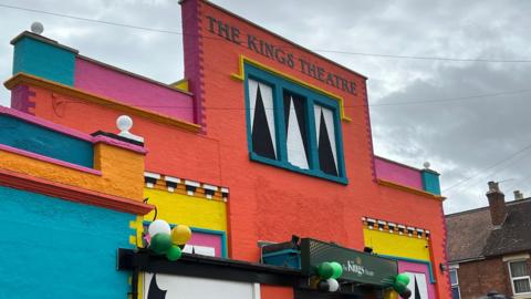 A building painted orange, blue, pink, yellow and with black and white accents. The words "The Kings Theatre" are printed at the top. Balloons are attached to the door.