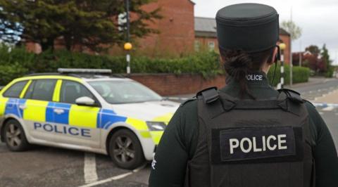 A police officer wearing a green uniform and a bullet proof vest with 'police' written on it. To the left of the officer is a yellow, white and blue police car.