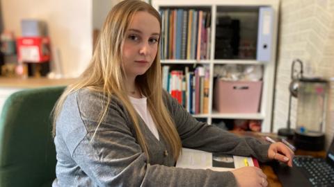 Amelia wearing a light grey top, sitting at a desk with a computer keyboard, with bookshelves in the background.
